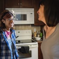 A trans teen with glasses, a blue flannel and brown skin smiles at their guardian while standing in the kitchen.
