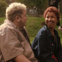 Two trans people with short curly hair smiling and looking at each other while sitting in the park.