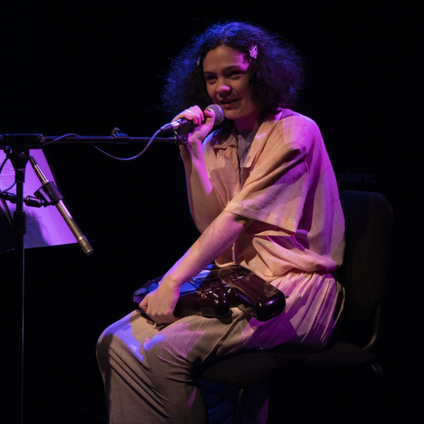Alex wears a soft pink top and bottom while sitting on a chair in front of a microphone. their hair is brown, curly, and clipped back with a pink barrett. The stage they are sitting on is black except for a purple light shining down on them.