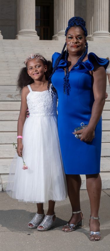 A Black woman wearing a blue dress with her arm around a Black child in a white dress